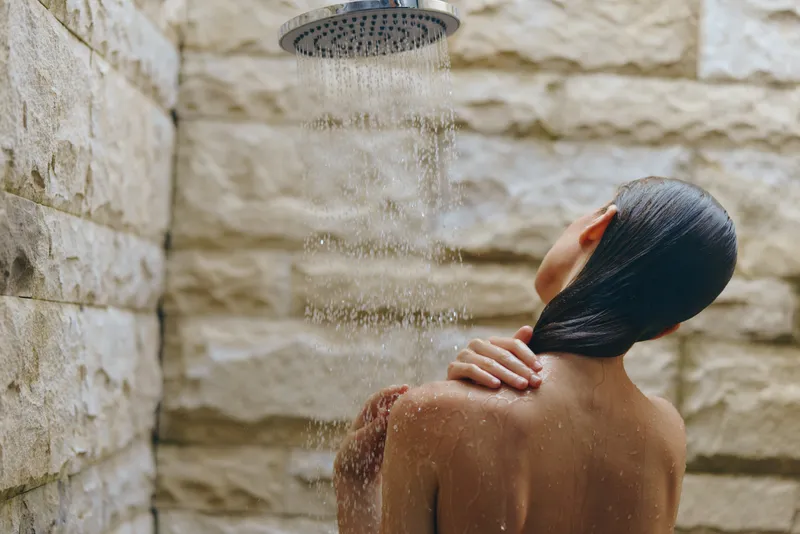 woman taking shower under rainfall showerhead in modern bathroom with stone walls, enjoying relaxation and self-care, wet hair, calm mood