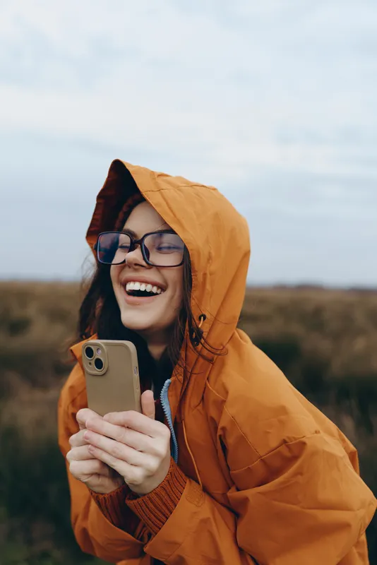 Cheerful smiling woman lifestyle outdoors, laughing in an orange hooded jacket, holding smartphone, sunlit autumn field nature backdrop, soft breeze, relaxed pose, open sky, daylight