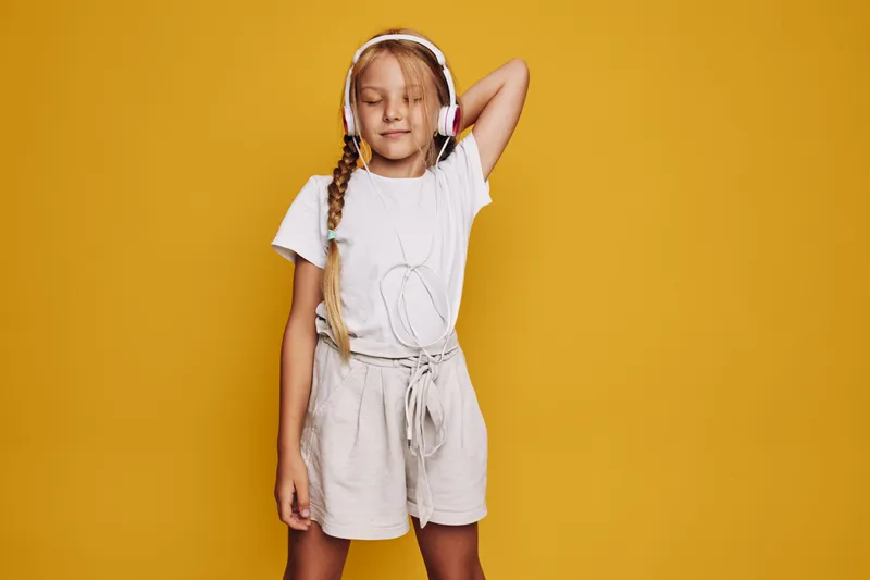 Young girl with long braided hair listens to music on headphones. She wears a white t-shirt and beige shorts against a bright yellow background. Concept for relaxation and enjoyment.