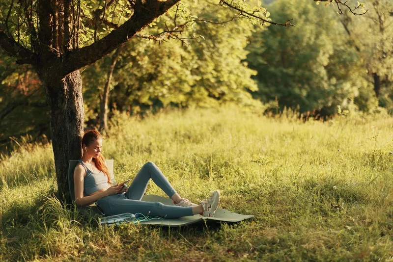 Relaxed young woman in casual athletic wear enjoying a moment under a tree, surrounded by nature, reflecting a healthy lifestyle and tranquility in the outdoors