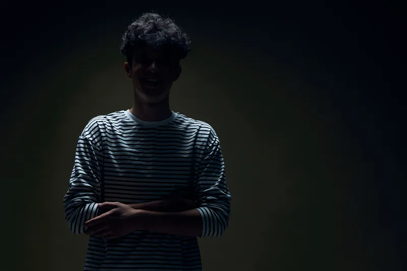 Silhouetted man with curly hair wearing a striped shirt stands with crossed arms against a dark background, creating a mysterious and moody atmosphere.