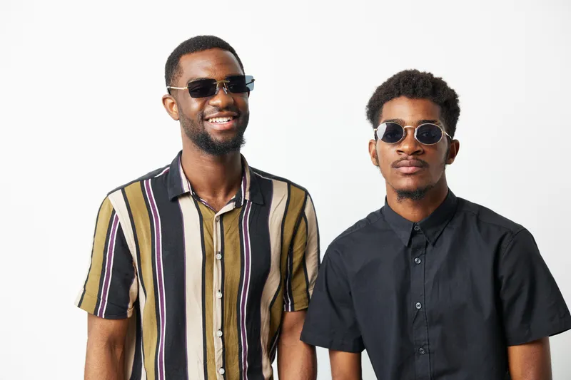 Two stylish black men wearing sunglasses, showcasing modern fashion and confidence. They smile warmly, creating an uplifting atmosphere. Perfect for lifestyle themes.