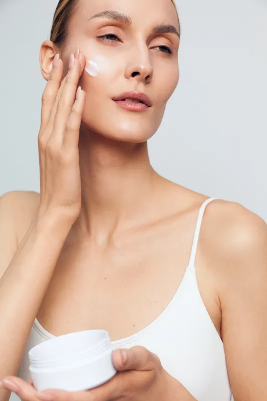A woman applies moisturizer in a bright, minimalist setting, capturing a calm skincare moment. Close up, soft light enhances healthy skin and everyday beauty.