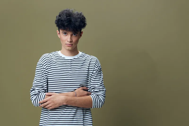 young man in striped shirt stands with crossed arms against green background, looking confident and relaxed, casual style, simple composition