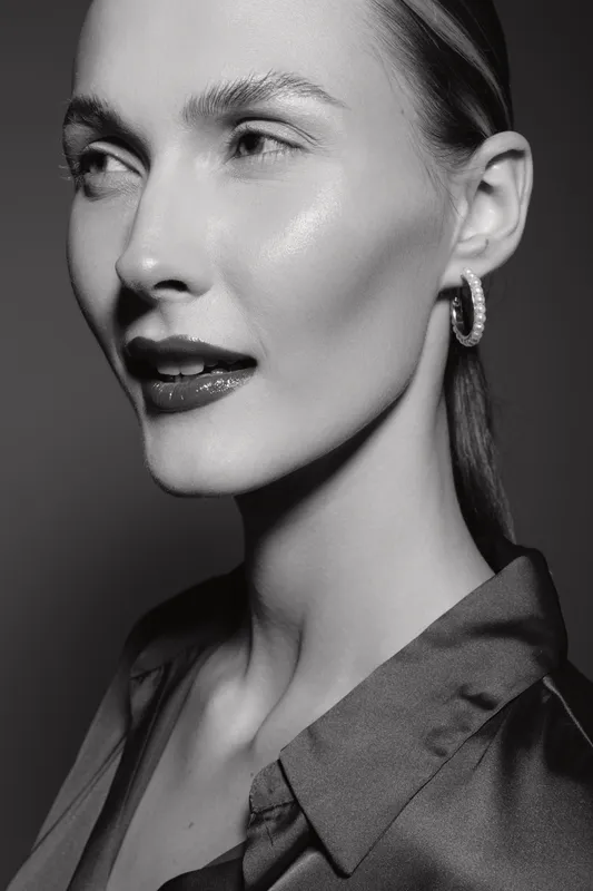 Close-up of a fair skinned Caucasian woman with slicked back hair wearing hoop earrings and dark lipstick, posing in studio with dramatic lighting and elegant expression.