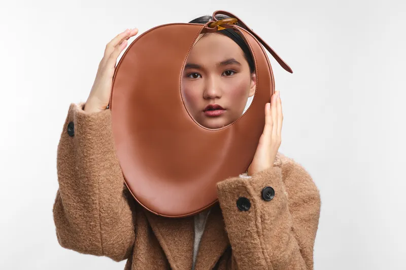 asian woman wearing a bold brown circular headpiece frames her face in a clean studio setting. she exudes calm confidence and playful curiosity, creating a modern, fashion driven atmosphere.