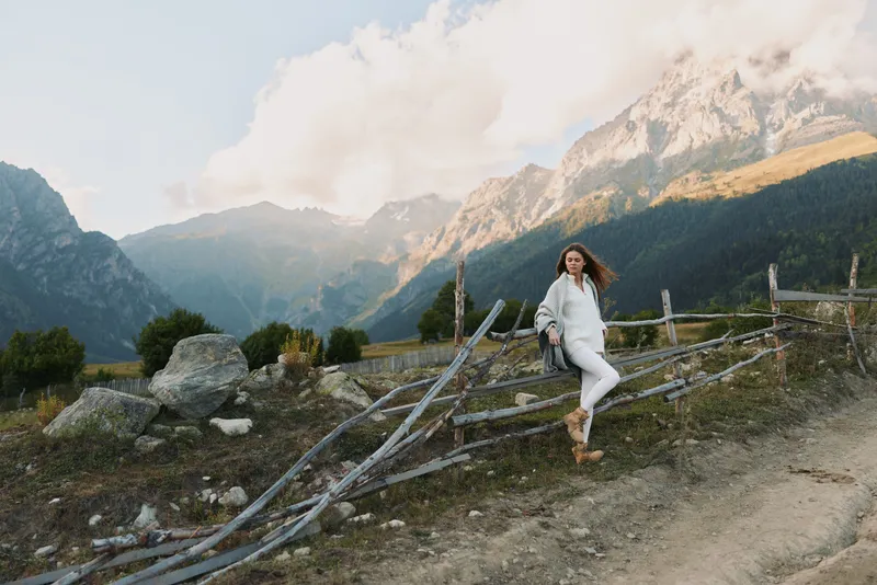 Young woman in white outfit leans on rustic wooden fence with mountain landscape in the background during a clear day.