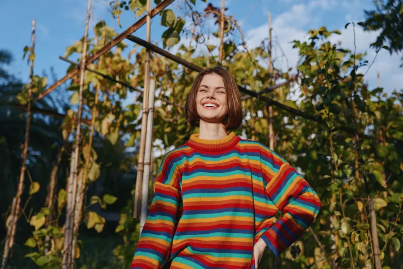 Woman smiling in a colorful striped sweater stands in a garden by a wooden trellis, short brown hair and casual outfit, confident pose and sunny lifestyle portrait for fashion wellbeing.