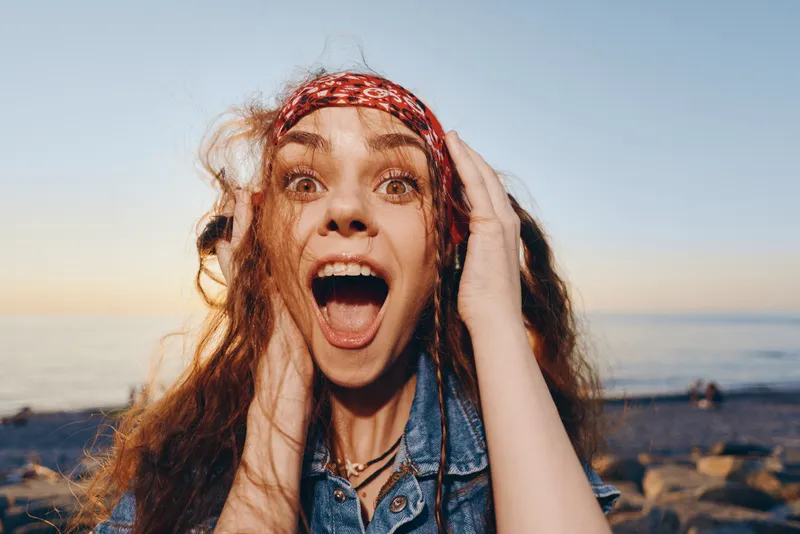 Excited woman with modern boho-western denim street style wearing a red headband by the seaside, capturing vibrant film color and joyful outdoor vibe.