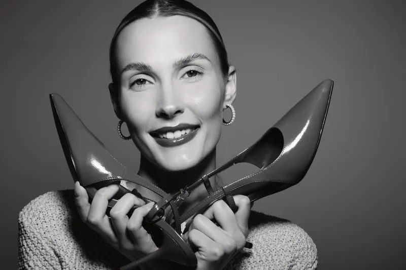 Smiling Caucasian woman with light skin holding pair of high heels near face isolated on grey background. Concept for fashion and footwear.
