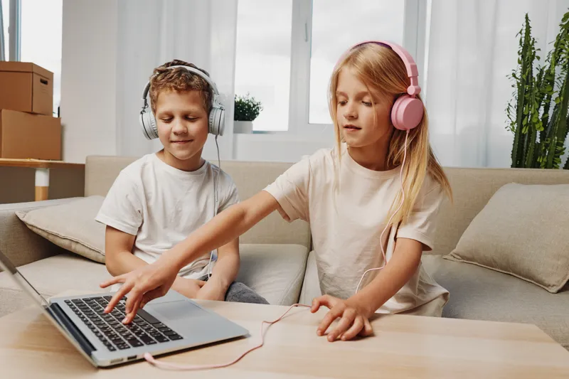 Two children with headphones use laptop together sitting on couch in bright living room. Concept of technology, learning, leisure, and digital entertainment for kids.
