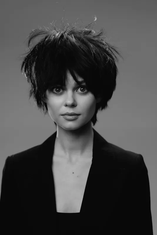 Black and white portrait of young woman with messy hair wearing dark blazer looking at camera.