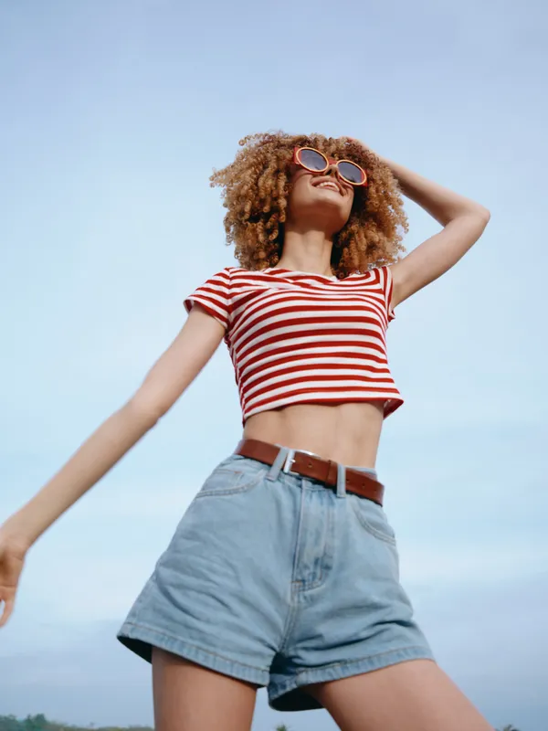 Young woman in a red and white striped crop top and high waisted denim shorts stands against a clear blue sky, radiating carefree summer energy and confident style.