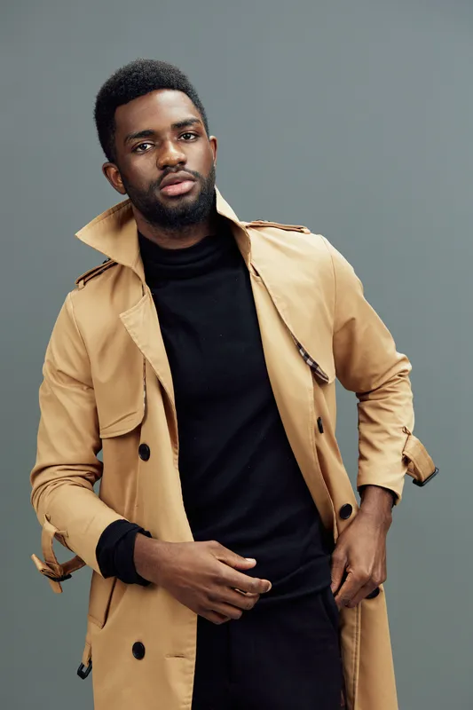 Stylish young man in a beige trench coat and black turtleneck posing confidently against a muted grey background, embodying urban fashion and sophistication