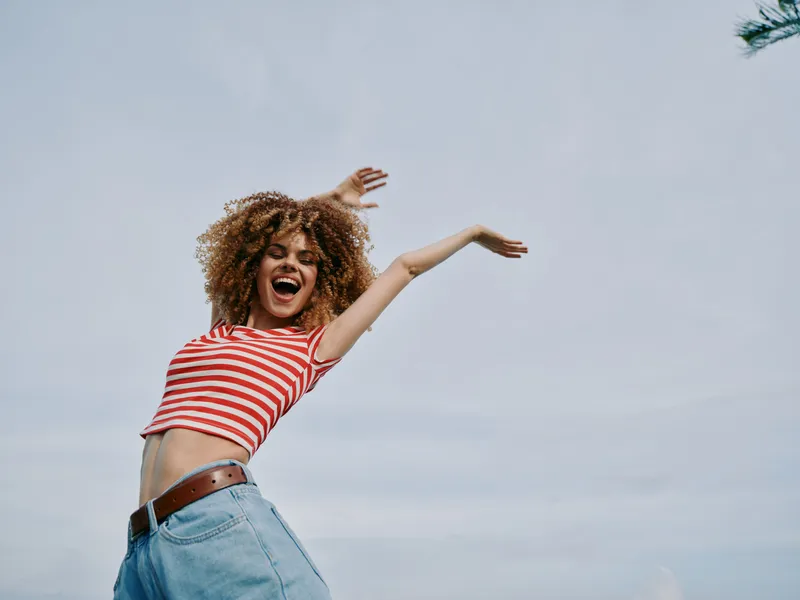Energetic young woman jumps with arms raised, wearing a red striped shirt and denim jeans, expressing carefree joy against a bright sky and gentle breeze.