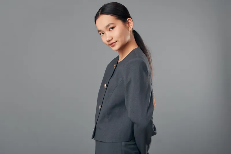 Young woman with long dark hair in professional gray suit standing sideways with calm expression on gray background for business portrait and confident fashion pose.