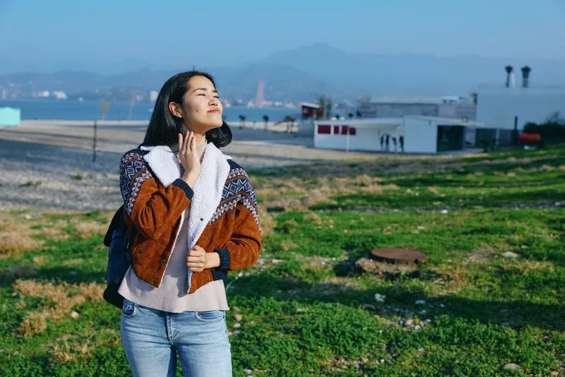 Woman enjoying outdoor scenery in a grassy field near the coast, wearing a brown coat and cozy scarf, with sunlit moments and a gentle breeze that enriches the peaceful open space