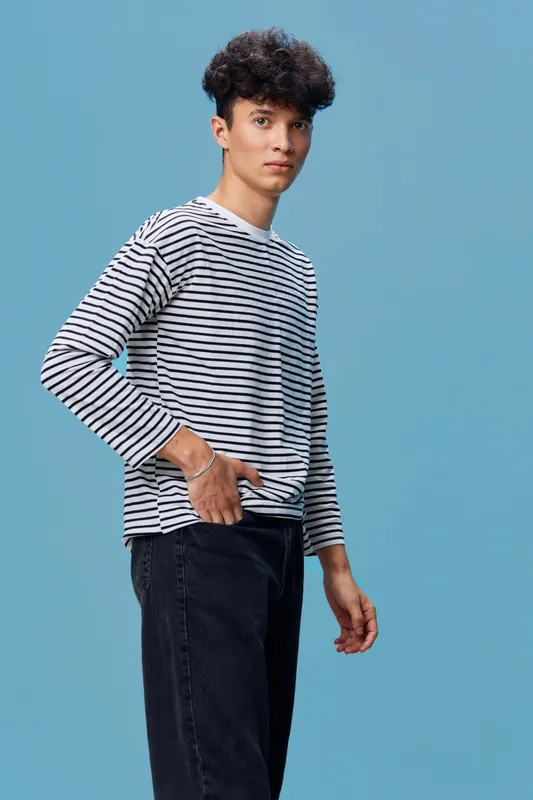 young man wearing striped long sleeve shirt and black jeans stands against blue background with thoughtful expression, casual style, curly hair, studio portrait