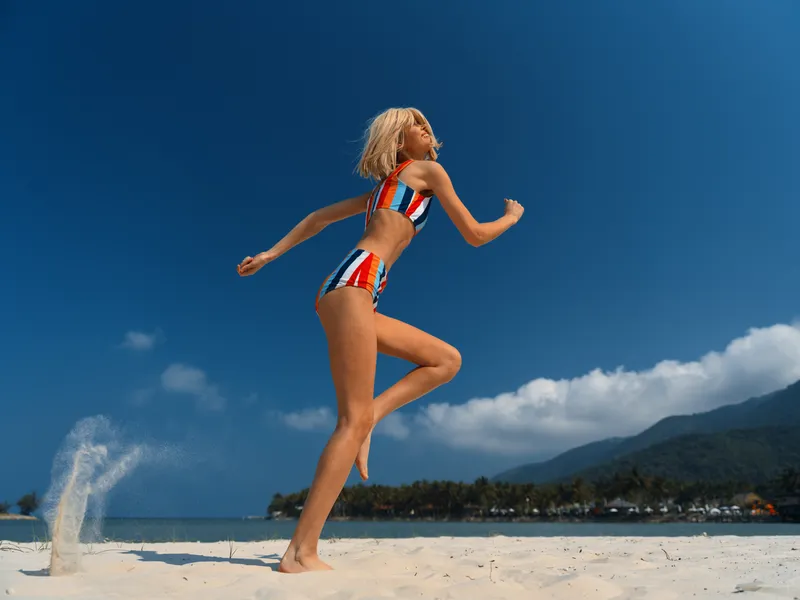 A young woman leaps midair on a sunlit beach, wearing a striped red, white, and blue bikini, captured against a clear blue sky and pristine white sand.