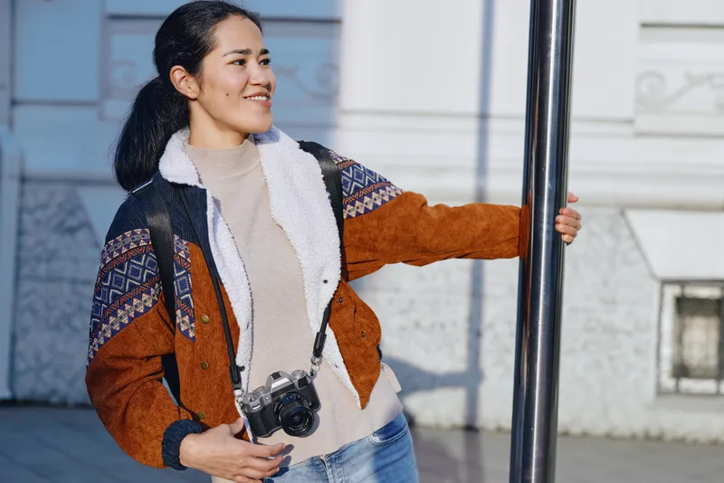 Woman with camera leaning against a pole in an urban outdoor setting, wearing a colorful jacket and jeans, smiling as she prepares for street photography and travel moments.