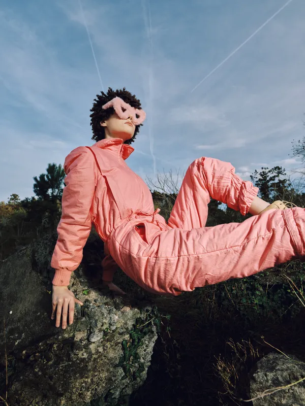 Puppet character wearing a pink jumpsuit and whimsical mask sunglasses lounges on a rock outdoors under a clear sky, creating a playful, quirky scene against natural scenery.