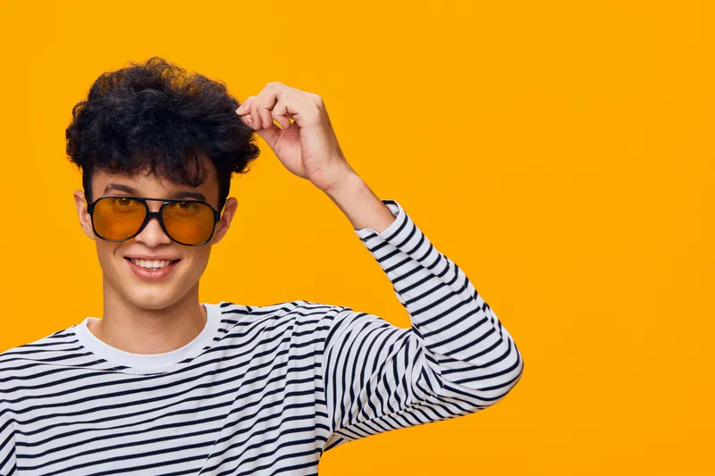 Young smiling man wearing striped shirt and yellow sunglasses poses against bright orange background, expressing confidence and casual style in vibrant studio setting.