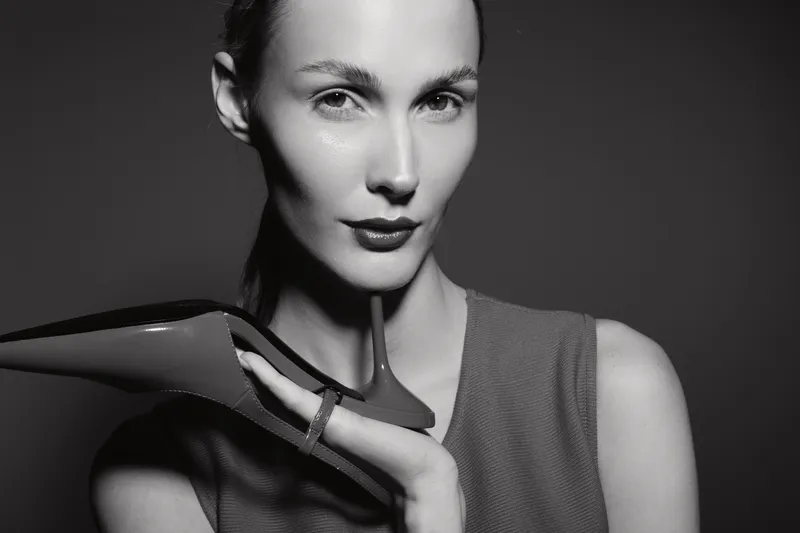 Portrait of a young Caucasian woman with pale skin holding a high heel shoe near her face in a studio. Concept of fashion, beauty, and elegance in black and white.