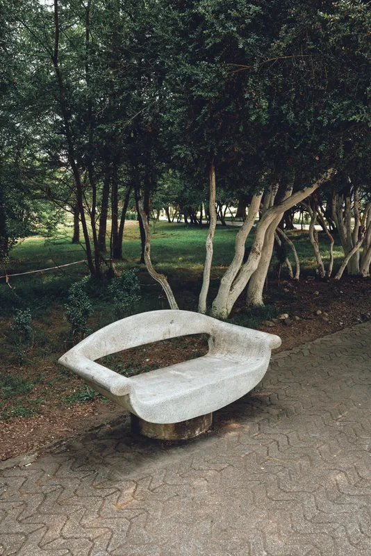 Stone bench in park under trees with curved seat by path, nature scene and outdoor rest