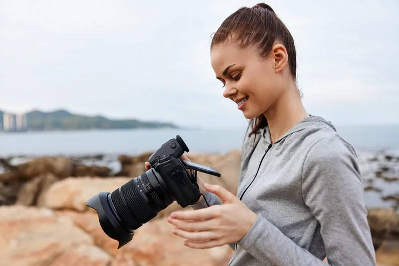 Young woman reviewing photos on her camera by the ocean, wearing a casual gray hoodie, showcasing joy and creativity in a serene coastal setting