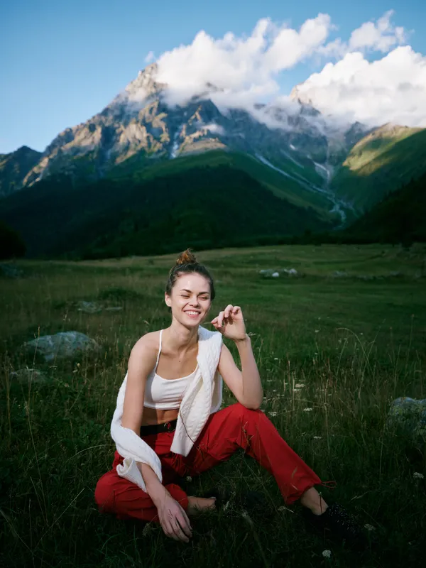 Outdoor mountain landscape with a smiling woman sitting on grass, casual outfit, towel draped over shoulder, capturing calm nature and scenic meadow atmosphere