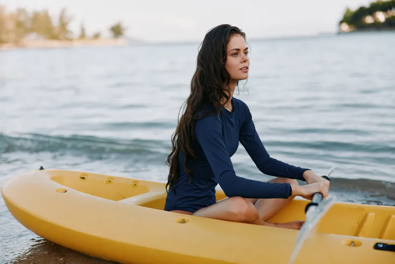 A woman sits in a yellow kayak on the shore, wearing a navy long sleeve shirt, gazing toward the horizon as gentle waves lap against sand and craft.