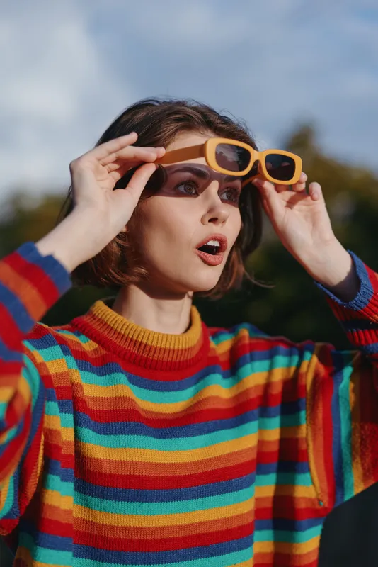 Woman in a colorful striped sweater lifts yellow sunglasses with a surprised expression, short brown hair, casual outfit and natural outdoor portrait for lifestyle and fashion.