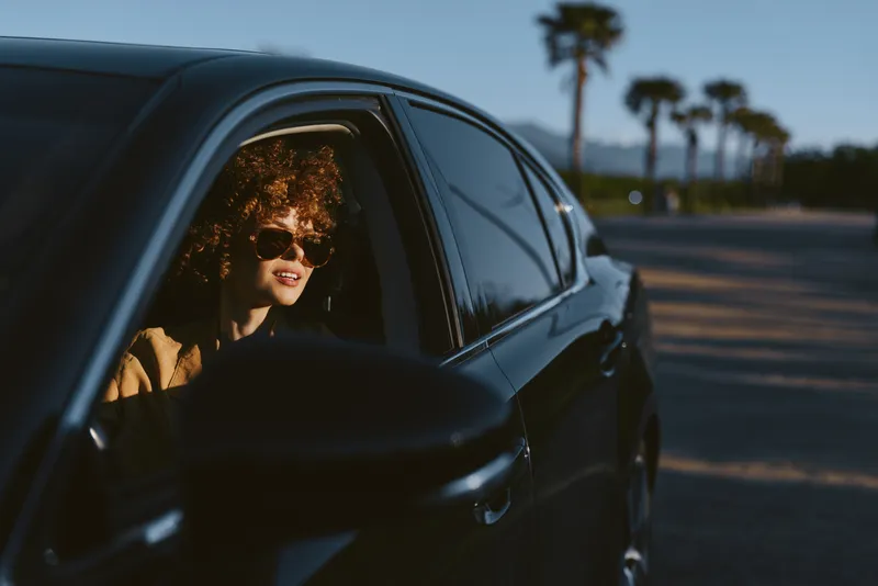 A woman with sunglasses drives a sleek car along a coastal road, shot in retro film color for a cinematic lifestyle moment. The warm light highlights her profile while palm trees line the horizon,