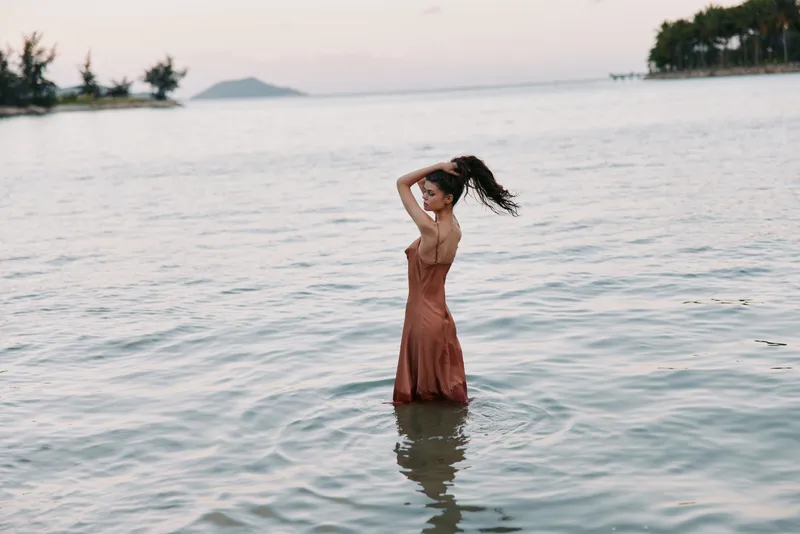 A solitary woman stands waist deep in calm sea water, rust dress flowing as she runs fingers through her hair, with soft sunset light and distant shoreline creating tranquility.