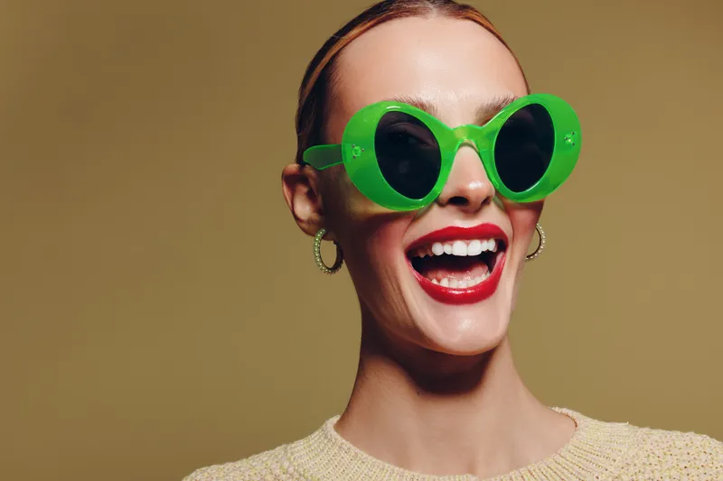 Smiling woman with bright red lipstick wearing large green sunglasses and hoop earrings on beige background. Concept of fashion, style, summer, and joyful mood.