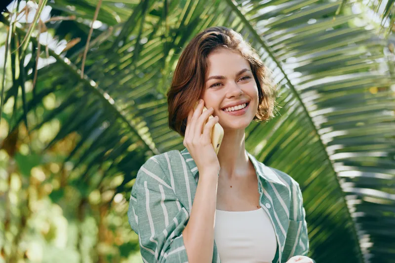 A cheerful woman speaks on a mobile phone outdoors, smiling warmly amid lush green palm leaves.