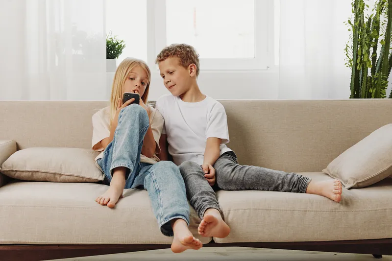 Two children sitting on beige sofa at home, a girl showing something on smartphone to a boy. Bright room with natural light and casual clothing, concept of sibling bonding and digital interaction.