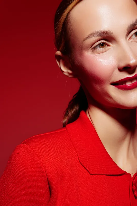 Close-up of a smiling woman with red lipstick and braided hair wearing a red sweater against a red background. Concept of beauty, fashion, and confident expression.