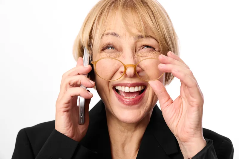 Smiling woman in studio, holding smartphone to ear, adjusting glasses with both hands, business communication, bright white background, closeup portrait, natural soft light