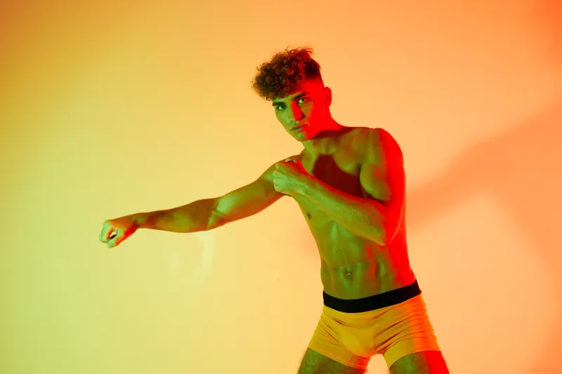 Young athletic man with curly hair practicing boxing punch in studio with colorful red and yellow lighting, fitness and workout concept for strength and training.