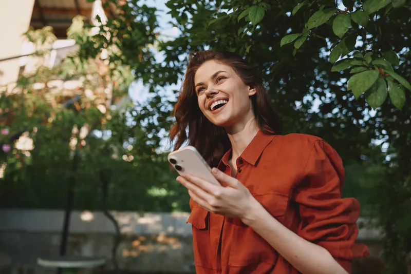 A smiling young woman wearing an orange shirt laughs while checking her smartphone in a sunlit outdoor garden
