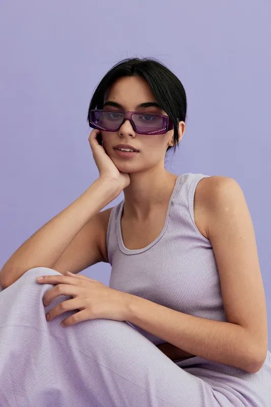 Young woman wearing purple sunglasses and a sleeveless lavender dress poses against a soft lilac background with relaxed, stylish expression and natural makeup.