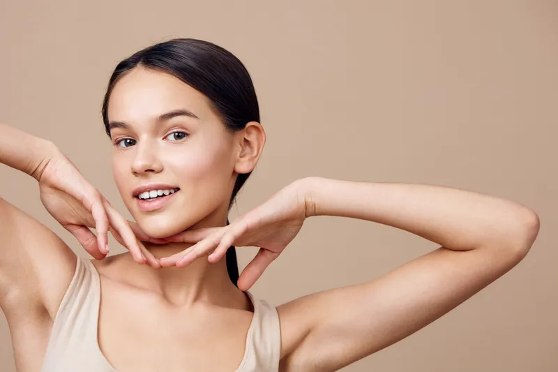 A young woman with a joyful expression poses with her hands under her chin, showcasing natural beauty against a soft beige background This image emphasizes elegance and self confidence