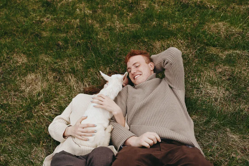 Relaxed man and goat kid lying on grass outdoors, sharing a warm, joyful moment in a sunlit pasture, embracing closeness and friendship, natural colors, gentle breeze, casual lifestyle scene