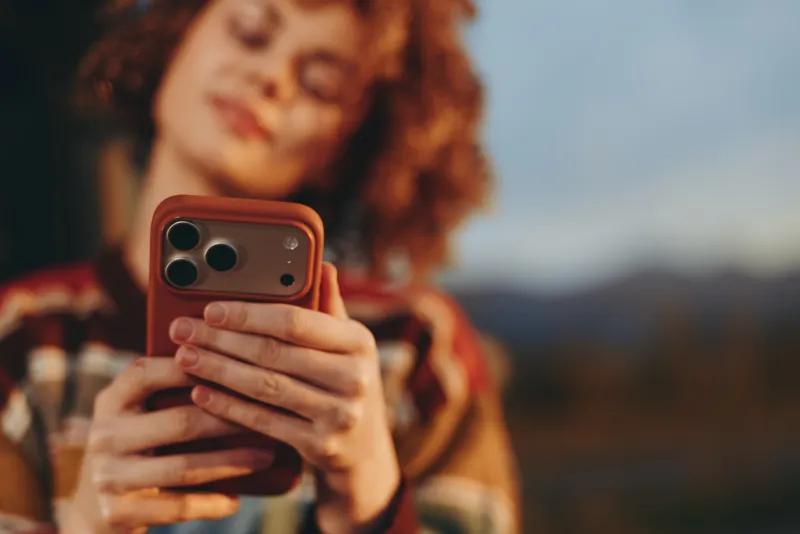 Woman with curly hair holding smartphone outdoors, lifestyle technology for communication, selfie moment in soft sunlight, shallow depth of field, blurred landscape background, relaxed casual