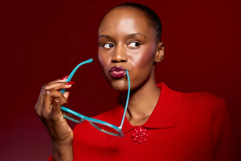 Portrait of dark skinned African woman in red sweater holding blue glasses near lips, looking sideways on red background. Concept of fashion, beauty, style, and confident expression.