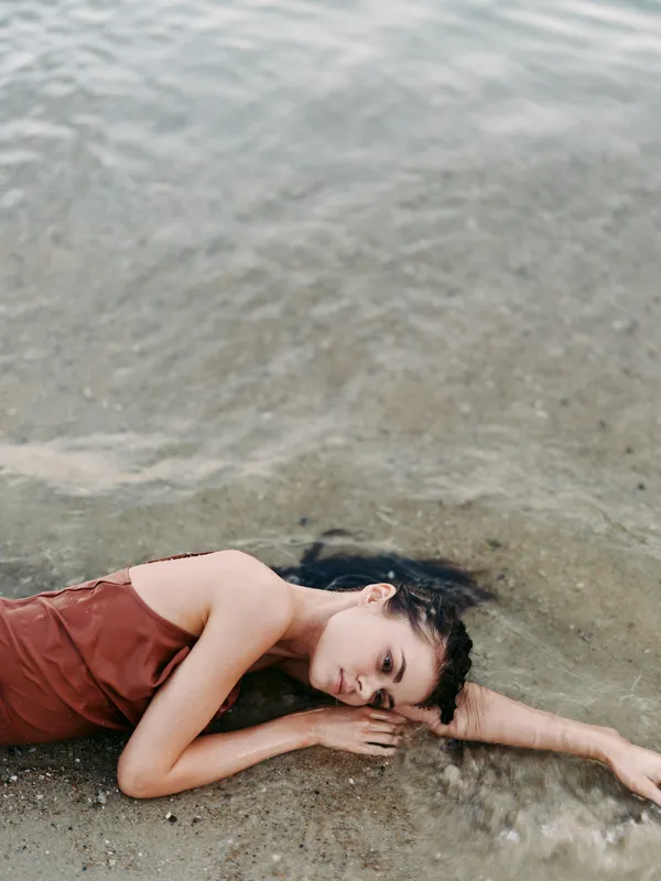 A solitary figure lies on damp sand beside gentle waves, wearing a rust colored dress, eyes closed, surrendering to the quiet rhythm of the shoreline.