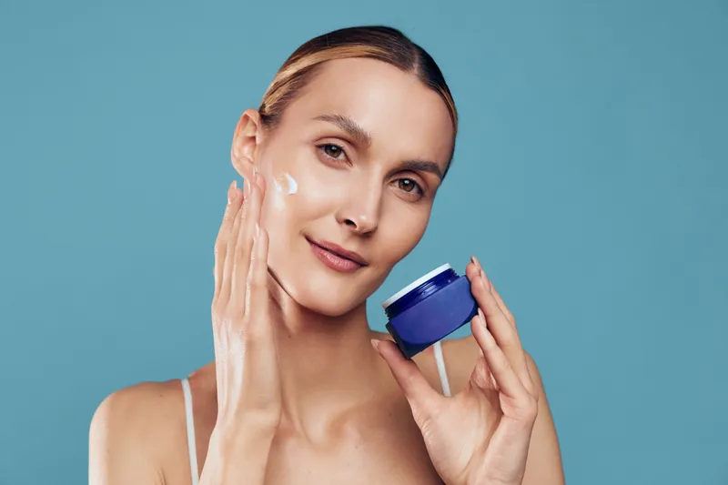 A young woman applies moisturizer cream on her face with one hand while holding a blue jar in the other. The clean skincare routine concept with a plain blue background.