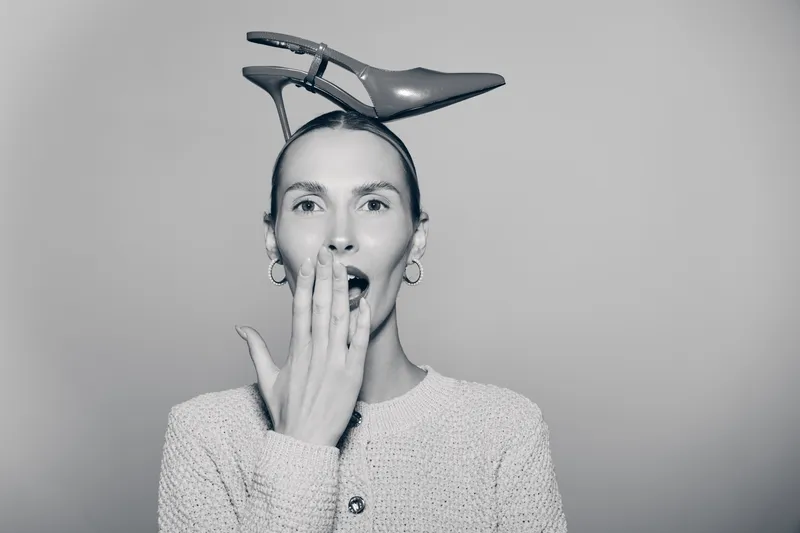 Surprised Caucasian woman with light skin holds high heel shoe on head and covers mouth with hand, concept of fashion and emotion.