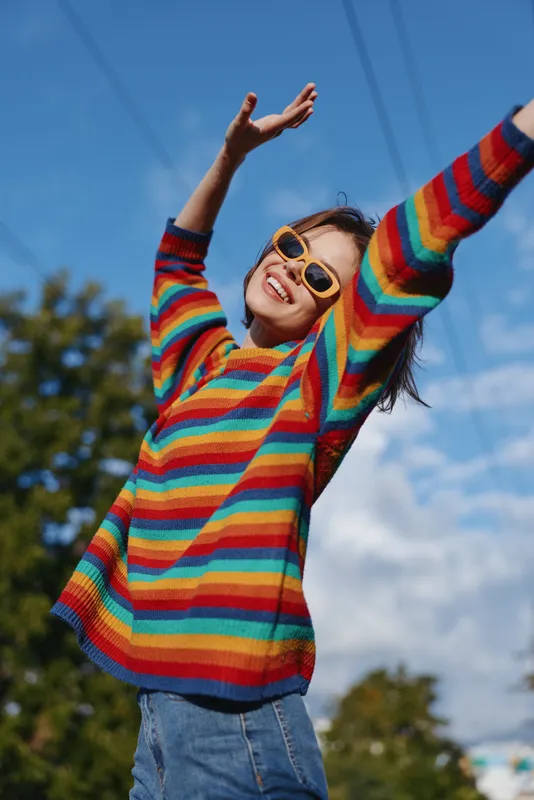 Woman in her mid 20s wearing a rainbow sweater and yellow sunglasses, smiling with arms raised outdoors under blue sky in a park, joyful casual lifestyle and fashion portrait.